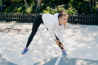 Koyu renk saçlı atletik Avrupalı bir kadının spor yaparken çekilen yatay çekimi ayak egzersizlerine eğiliyor açık havada giyinirken gülümsemeler takıyor trenleri ısıtıyor kasları tam boy gösteriyor.