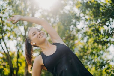 Atkuyruklu neşeli sporcu kadın, dışarıda ağaçlara karşı egzersiz yapıyor güneşli bir günün tadını çıkarıyor, kablosuz kulaklıkla müzik dinliyor, sabah antrenmanları yapıyor, kardiyo egzersizlerini ısıtıyor.