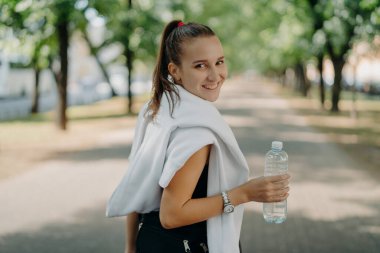 Parktaki kardiyo çalışmasından sonra, atkuyruklu neşeli, mutlu, sportif bir kadının arkasından bakınca yorucu sabah koşusu düzenli olarak spora gittikten sonra bir şişe temiz su tutuyor.