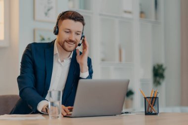 Mutlu iş adamı bilgisayarı ve klavyeyi kullanarak masada oturup kulaklıkla video görüşmesi yaparak web konferansına katılıyor. Çevrimiçi iş kavramı