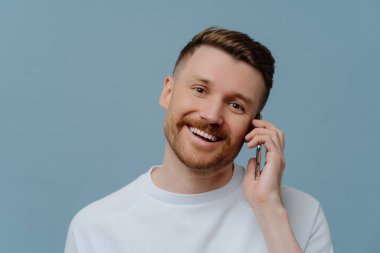 Kapalı alanda çekilen yakışıklı sakallı adam, mavi arka planda izole edilmiş beyaz tişört giymiş diş tebessümleri ile telefonda konuşmaktan hoşlanıyor. Traşsız mutlu adam arkadaşlarıyla haberleri tartışıyor. İletişim