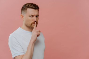 Tişörtlü, sessiz ve sessiz bir jest yapan genç ve ciddi bir adam. Parmağını ağzının yakınında tutuyor ve pembe stüdyo arkaplanına yaslanırken kameraya bakıyor. Mesaj için boşluk kopyalıyor.