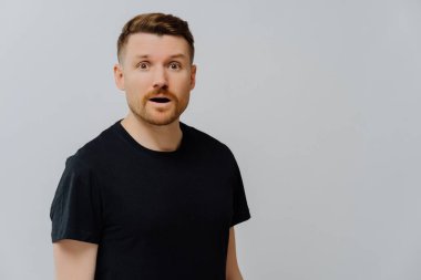 Stüdyoda, siyah tişörtlü, şaşkın bir şekilde kameraya bakarken gri arka plan üzerinde duran, şaşırmış ve şaşırmış yakışıklı kızıl adamın görüntüsü. Şaşırtıcı duygu.