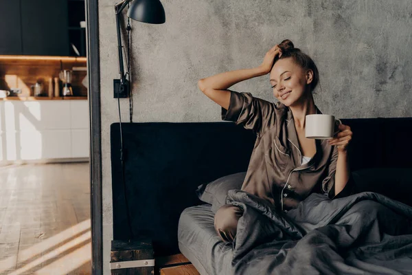Young relaxed woman in stylish home suit waking up with cup of coffee in bed in cozy stylish bedroom at home, stretching from night sleep and enjoying morning leisure time and sunshine from window