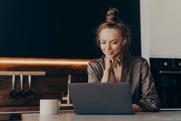 Young female freelance worker in pajama drinks coffee in morning for successful productive working start while sitting in kitchen with laptop and checking emails. Remote work and freelance concept
