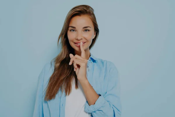 Young pretty woman wide eyed with smile asking for silence or secrecy with finger on lips with hush hand gesture isolated on blue background. Positive emotions and facial expression, body language