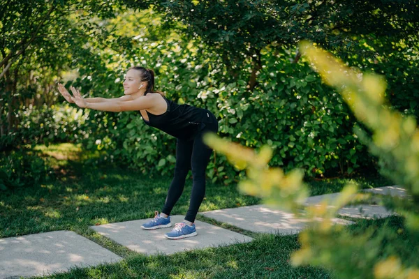 Energetic young woman with pony tail has workout outdoors leans forward stretches arms exercises before running warms up body wears sportsclothes poses against green trees in garden or park.