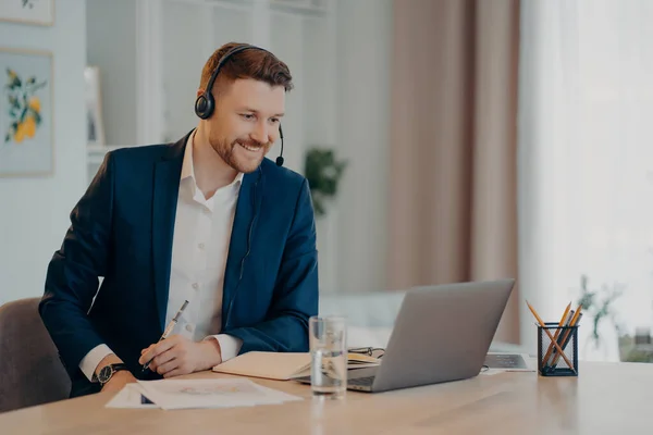 Smiling glad successful businessman practices online conference has distance meeting with colleagues wears headset focused at laptop computer wears formal clothes. Freelance working concept.