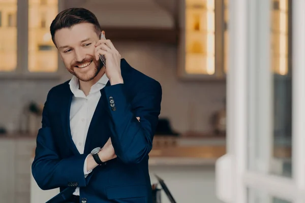 stock image Positive successful businessman in suit standing in living room at home and enjoying talk on mobile phone with partner, holding smartphone and talking about job. Business communication concept