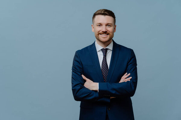 Waist up shot of self confident bearded male entrepreneur keeps arms crossed smiles gladfully dressed in formal clothes isolated over blue studio background copy space for your advertising content