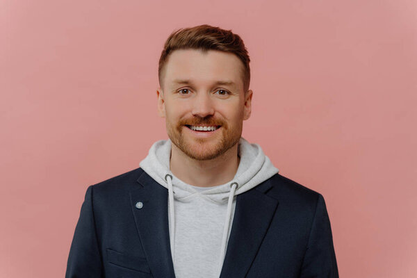 Portrait of positive attractive red haired guy wearing hoodie and jacket smiling at camera, young good looking cheerful man with beard posing in stylish casual clothes on pink studio background