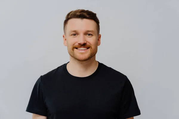 stock image Handsome good looking ginger unshaven man smiling happily at camera while standing in casual black shirt, being positive and friendly, looking with cheerful expression while posing over grey wall