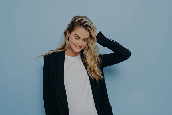 Beautiful smiling female with blonde dyed hair feeling happy, wearing black blazer over white tshirt while standing isolated against blue background, positive woman keeping eyes closed and smiling