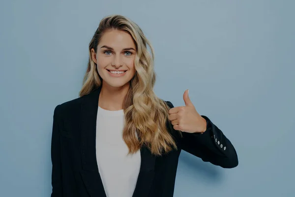 Satisfied blonde woman in formal outfit showing thumbs up gesture, giving positive feedback, demonstrating approving expression while looking at camera, standing alone next to blue background