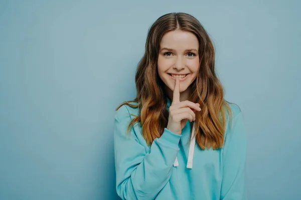 Attractive young teen girl smiling while touching lips with pointer finger showing shushing sign wearing casual comfortable sweater posing isolated on blue studio background