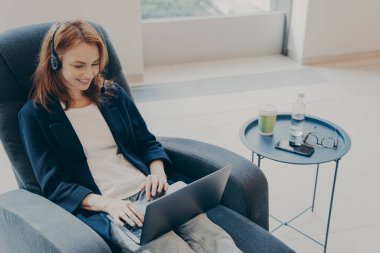 Kulaklık takmış genç girişimci bayan partneriyle online sohbet ediyor, evde koltukta otururken dizüstü bilgisayar kullanıyor, bilgisayardaki video görüşmesinin keyfini çıkarıyor. Uzaktan öğrenme ve çalışma kavramı