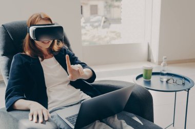 VR kulaklıklı genç mutlu iş kadını bir şeylere dokunmaya çalışıyor, iş için sanal gerçekliği kullanıyor, rahat evinde oturuyor, uzaktan çalışırken kadın serbest yazar gözlüklerini test ediyor.