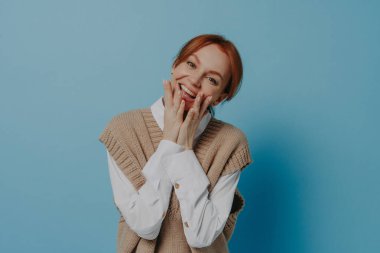 Mutluluk. Kameraya gülümseyen, ağzı iki eliyle kaplayan ve mavi arka planda izole bir şekilde dikilirken mutlu hisseden aşırı neşeli, genç kızıl saçlı kadının stüdyo portresi.