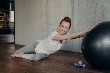 Spor giyim sektöründe aktif ve sağlıklı bir kadın ahşap zeminde uzanmış, büyük gümüş bir spor topu ile kameraya gülümsüyor ve spor salonunda egzersiz yapıyor. Spor ve sağlık hizmetleri