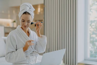 Genç Avrupalı bir kadının fotoğrafı, dizüstü bilgisayarda yoğunlaşmış makyaj fırçasıyla yüz pudrası uyguluyor. Kendi güzellik blogunda, iç mekanlara karşı rahat bornozla poz veriyor.