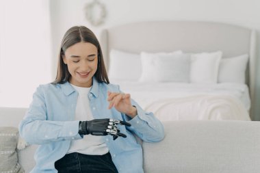 Happy girl is setting her electronic bionic limb. Attractive european woman with artificial arm at home. Smiling woman has modern cyber prosthesis. Engineering and innovation concept.
