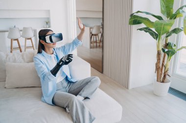 Engelli kişi rehabilitasyona ve siber ortamda oynamaya gidiyor. Oturma odasında kanepede oturan beyaz bir kadın. VR gözlüklü engelli kız. Biyonik uzuv protezli ampüte.