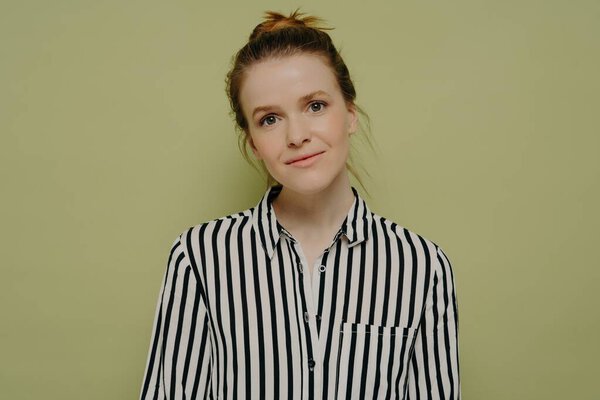 Portrait of calm young european woman with hair in bun wearing black and white striped shirt looking at camera with tilted head and slight smile while standing alone next to green wall