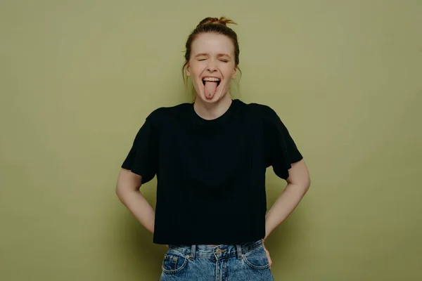 Crazy young woman wearing casual clothes with hair in bun making a goofy face with wide open mouth, showing her tongue and having fun while standing isolated next to green background