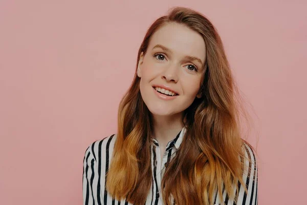 stock image Attractive young female with wavy medium length ginger hair in stripy black and white blouse tilting head while posing over light pink background. Human emotions concept