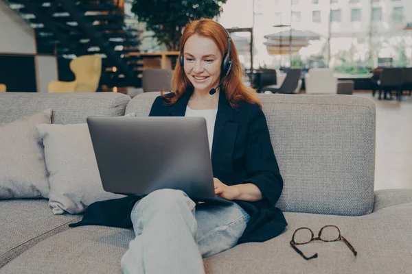無線ヘッドセットを身に着けている赤い髪を持つ笑顔の白人女性フリーランサー ラップトップ上のビデオ通話を介してクライアントと通信します オンライン言語のレッスンを与えます 地元のコーヒーショップで居心地の良いソファに座って — ストック写真