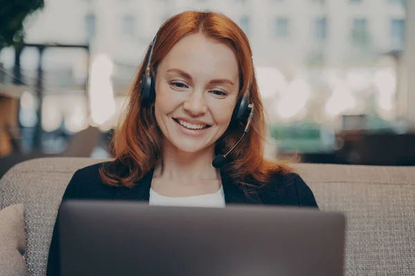現代のオフィスでソファに座っている間 ノートパソコンで同僚とオンラインで話してヘッドセットを着て笑顔の若い赤毛の女性は 自宅からのリモートワーク中にオンライン会議に参加 — ストック写真