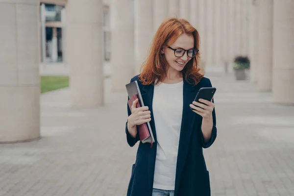 Young positive red haired business woman in elegant outfit using mobile phone, texting sms or checking email while standing outdoors after hard working dy, holding laptop and note book