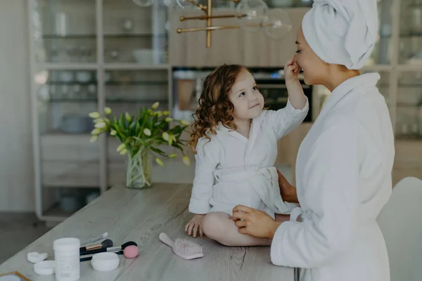 Pretty curly small girl touches nose of her mother dressed in white soft dressing gown sits on table with cosmetic products going to do makeup for mom pose together against cozy home interior