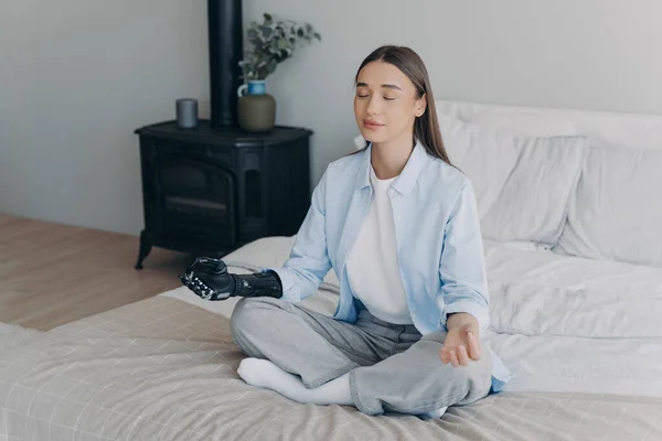 Young handicapped woman relaxing in cosy room. Girl practicing yoga in lotus pose on bed in her bedroom. Stress relief at home. Attractive european girl has cyber prosthesis. Modern artificial limb.