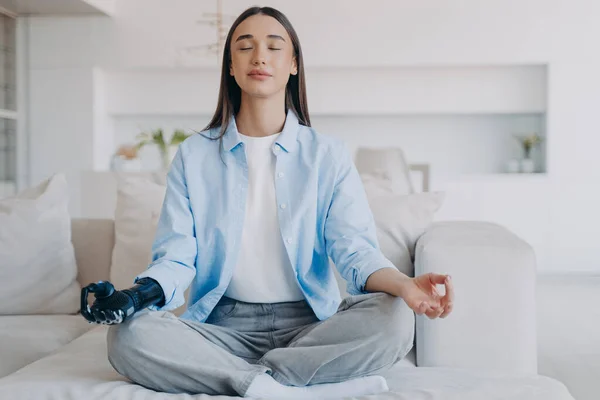 Serene disabled woman is sitting in lotus pose on couch. Lady practicing morning yoga with her eyes closed. Attractive european girl has myoelectric cyber prosthesis. Modern artificial limb.
