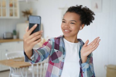 Young mixed race teen girl wear wireless earphones holds smartphone use video call, greet friend, waving hand. Friendly biracial female blogger start talk in social network, welcoming followers.