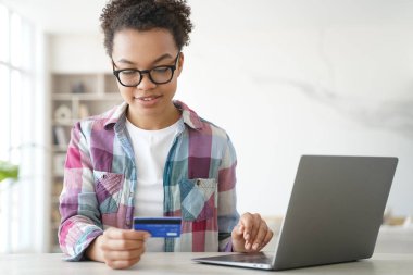 Afrikalı Amerikalı genç kız kredi kartı alışverişini dizüstü bilgisayardan yapıyor. Çift ırklı genç bir kadın evdeki bilgisayardan ödeme alıyor, bankacılık hizmetlerine göz atıyor, ikramiyeyi kontrol ediyor..
