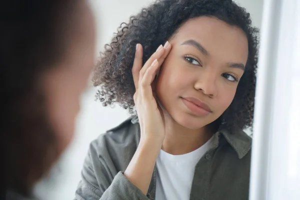 stock image Morning skincare. Confident afro girl is applying cream to her face and looking into mirror. Skin cleansing and moisturizing with lotion. Daily beauty routine and femininity of teenage girl.