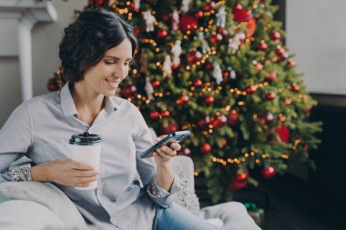 Gülümseyen Avrupalı kadın evde yeni yılın tadını çıkarıyor, cep telefonu ve kahve fincanı olan mutlu İspanyol kadın Noel ağacının yanında oturuyor, akıllı telefon kullanıyor, tatil dileğiyle mesaj yazıyor.