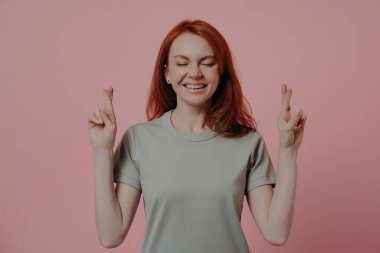 Umut dolu kızıl saçlı, sıradan gözleri kapalı bir bayan öğrencinin stüdyo fotoğrafı. Yüzünde batıl inançlar olan şaşı parmaklar ile sınavdan önce iyi şanslar diliyor. Pembe arka planda izole edilmiş.