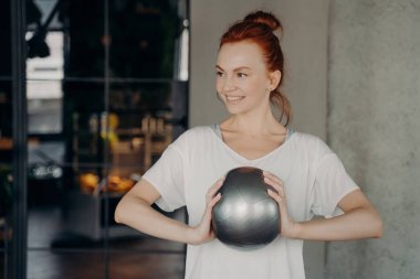 Sportif, kızıl saçlı, spor kıyafetleri giyen, elinde iki eliyle küçük bir fitball topu tutan ve spor salonunda çalışmaya hazır birine gülümseyen güzel bir kadın. Sağlık ve kişisel bakım kavramı