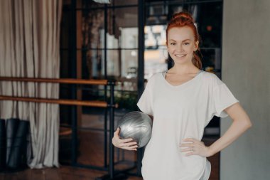 Beyaz tişörtlü, güzel gülüşlü, bir elinde gümüş renkli spor topu olan ve pilates derslerinin başlamasını bekleyen genç, mutlu bir zencefil fitness eğitmeni. Sağlıklı yaşam tarzı ve kapalı alanda spor