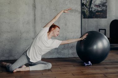 Aktif kızıl saçlı fitness kızı spor yaparken parke zeminde, pilates stüdyosundaki beton gri duvarın yanında çıplak oturuyordu. Etkin yaşam tarzı ve vücut bakımı kavramı