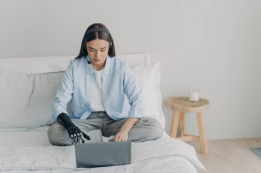 Disabled european woman with artificial arm working on laptop in bedroom. Freelancer has online meeting sitting on her bed using headset and microphone. Handicapped girl is remote assistant.