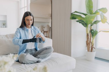 Disabled european girl is getting used to bionic arm. Cyber sensor hand has processor chip and buttons. Female amputee is sitting and adjusting electronic artificial arm at home. Carbon robotic hand.