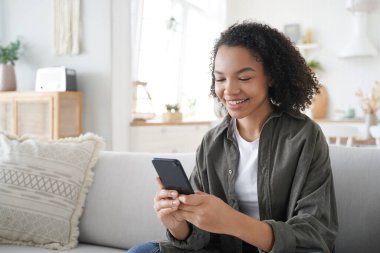 Curly african american girl has fun with phone at home. Happy hispanic woman is browsing social media and smiling. Chatting with friends remotely. Messaging with classmates. Communication concept.