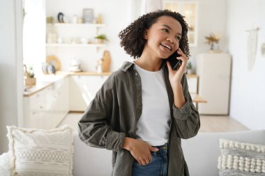 Modern african american young female answer call, talking on phone with a friend at home on sunny day. Biracial teen girl enjoying conversation by smartphone discussing positive good news on weekend.