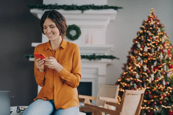 Joyful european woman using cellphone while sitting home office with Christmas tree on background, smiling hispanic female checking email or news feed of social network account during xmas holidays