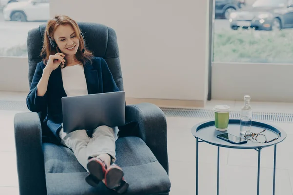 同僚とオンラインで話す成功した赤毛の女性起業家は 開いたラップトップの前でリラックスしたポーズでアームチェアに座って 自宅で遠く働いているヘッドセットを使用しています フリーランスのコンセプト — ストック写真