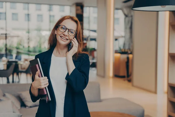 現代のコワーキングスペースに立ち ノートパソコンを持ちながら 職場でビジネスコールをする美しい赤髪のビジネスマンの笑顔 モバイルでアイウェアコンサルティングクライアントの幸せな女性 — ストック写真